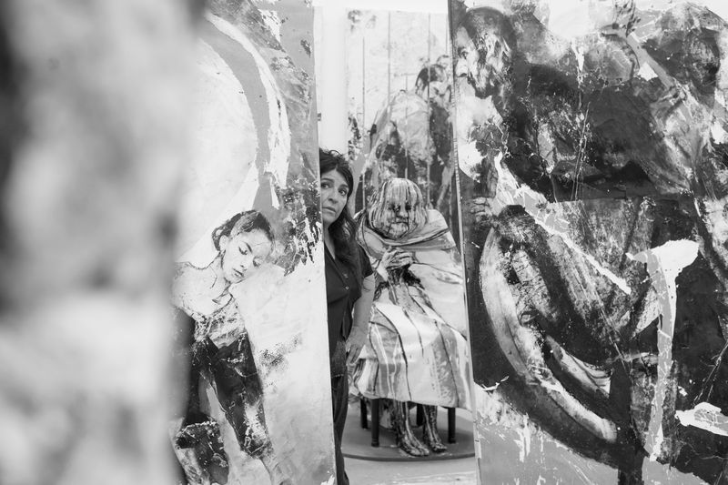 Une artiste se tenant derrière des toiles exposées dans une galerie d'art, avec des peintures expressives en noir et blanc en arrière-plan.